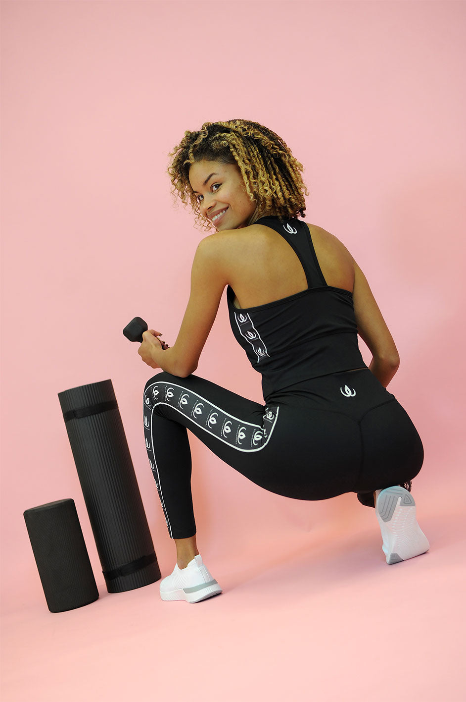 La fascia con logo corre lungo il lato quando la modella si gira di spalle, con manubrio e foam roller accanto.