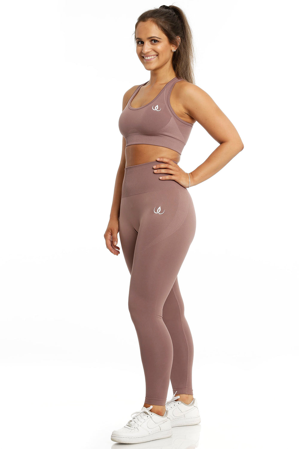 Sports bra and leggings in the same rose brown tone. The model stands with one hand on her hip, in white shoes.