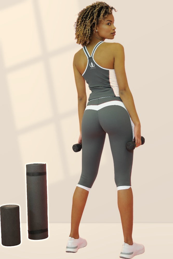 Grey capri tights with white edging at the knees, worn with a grey vest. The model is holding two dumbbells.