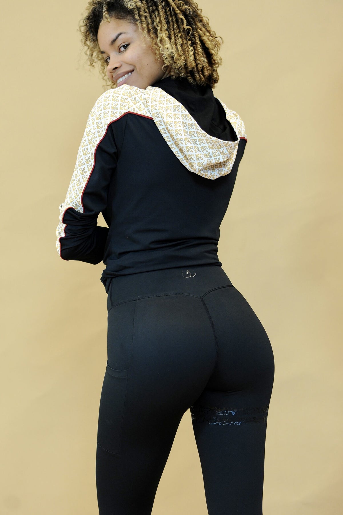 Sopra la spalla si vedono il cappuccio con motivo oro e il profilo rosso della giacca. La modella indossa leggings neri.