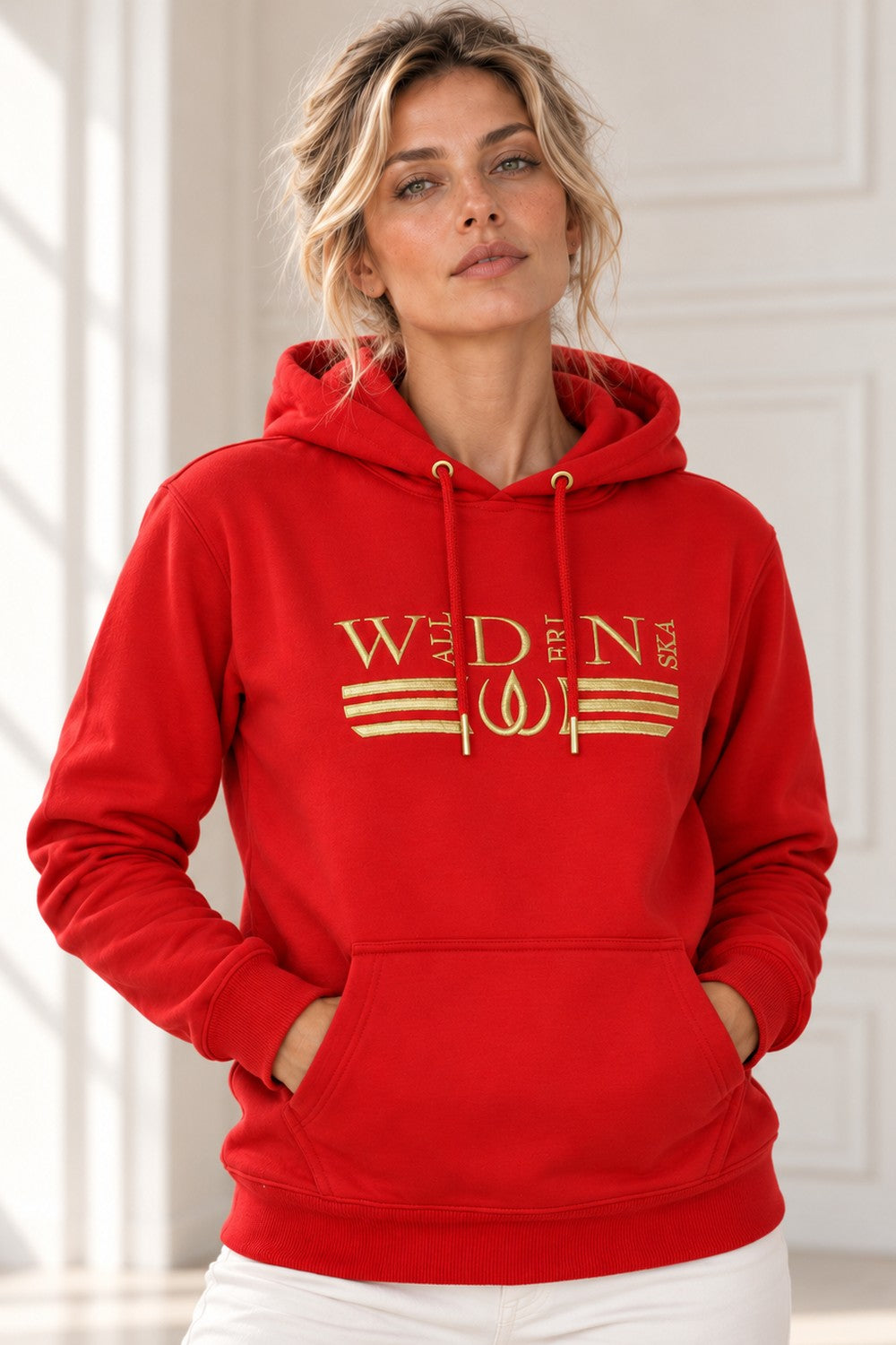 Röd Deluxe Hoodie med guldbroderad logga och guldränder över bröstet, buren till vita byxor.