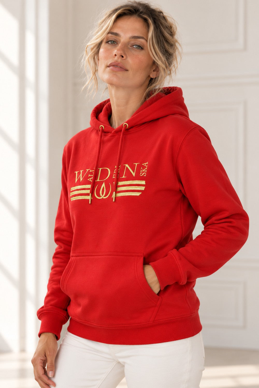 Modellen i den röda huvtröjan med guldbroderi och känguruficka, i en ljus studio med fönsterljus.