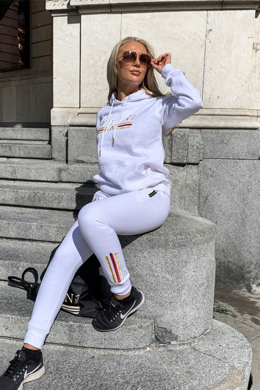On stone steps in town, the model wears white joggers with a gold stripe at the ankle and a white hoodie.