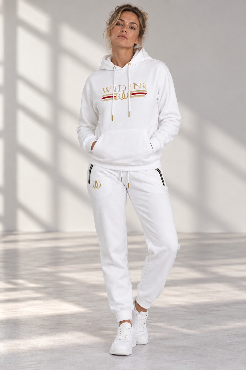 Modellen i det vita Deluxe-setet, hoodie och joggers med dragkedjor vid fickorna och guldlogga.