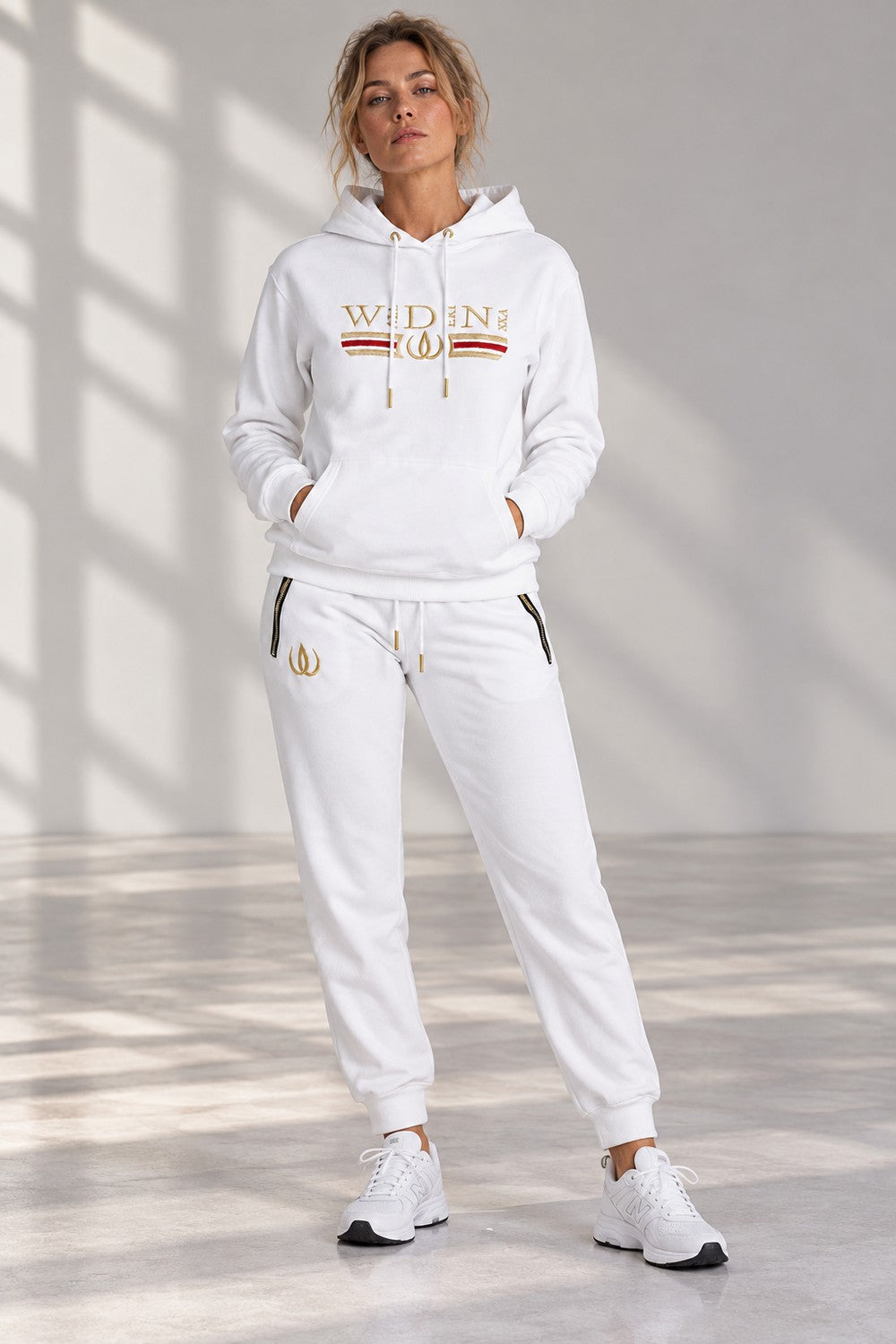 Vit Deluxe Hoodie och Deluxe Joggers Vita i helfigur, med guldlogga på låret och vita sneakers.