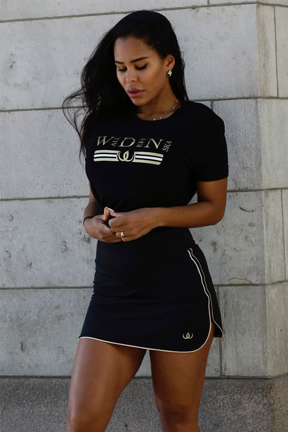 The model stands against a light wall in the black skirt and a black t-shirt with gold print.