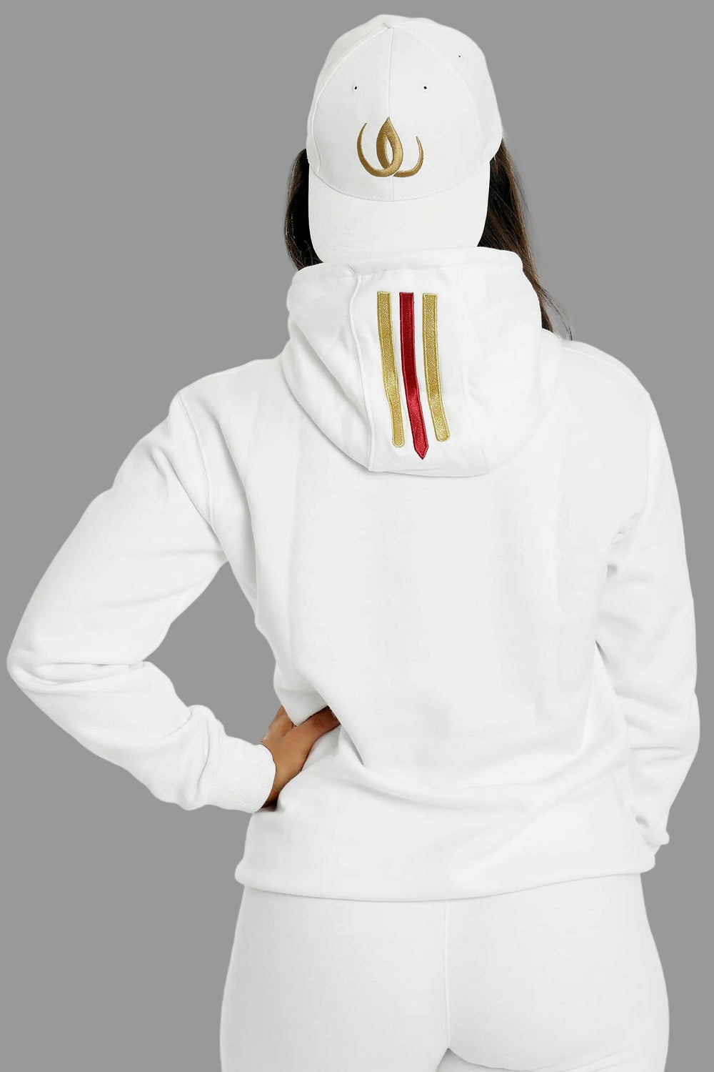 Back of the white hoodie with two gold stripes and one red on the hood. The model wears a white cap.