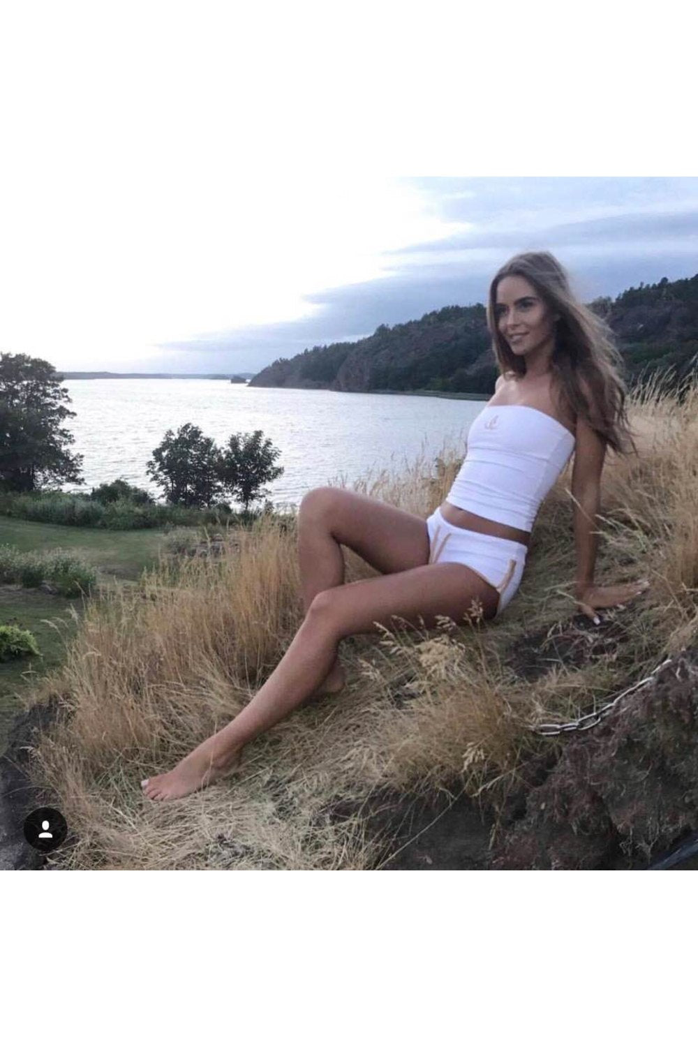 The model leans back in the grass by the water, in white shorts with gold edging and a white bandeau top.