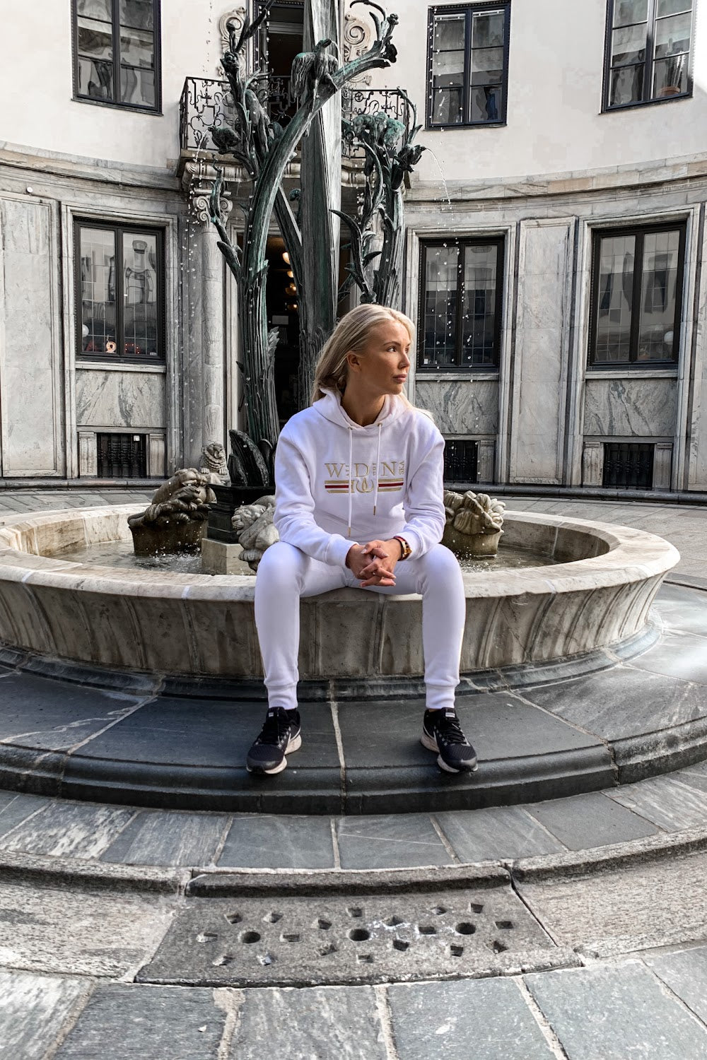 On the edge of the fountain the model sits in the white hoodie, with a statue in the background.