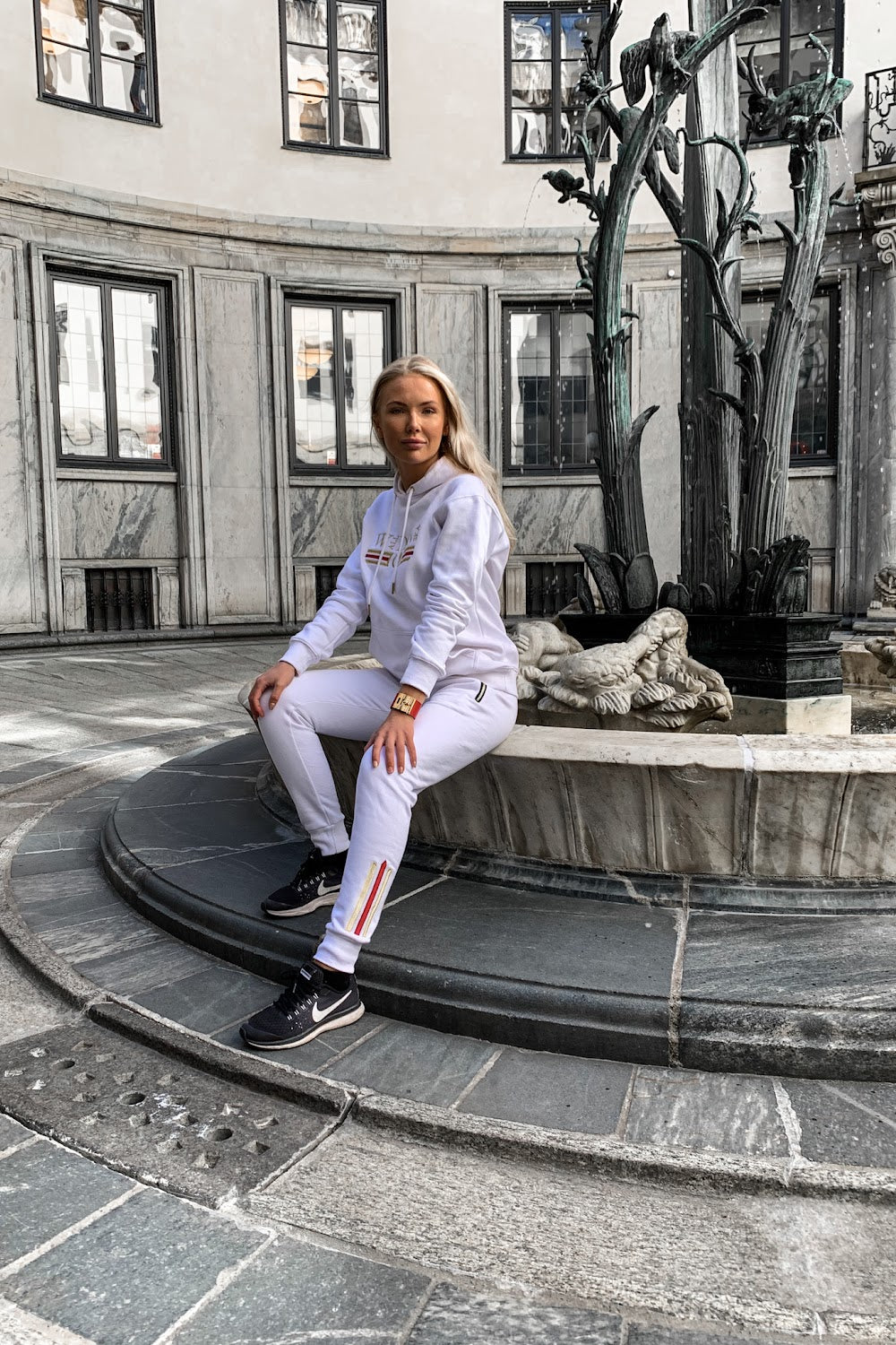 On the edge of the fountain in a seated position, the gold stripe along the leg of the white joggers is visible.
