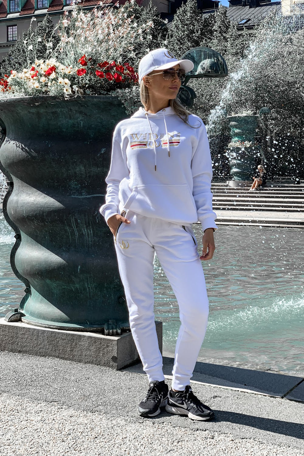 By a dark urn the model stands with hands in the pockets, in a white hoodie and white joggers.
