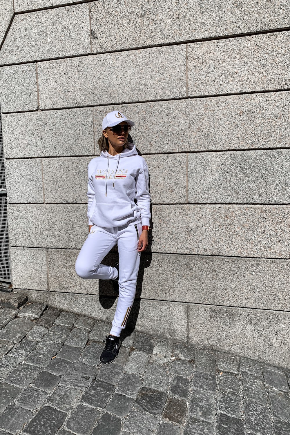 Against the stone wall the model leans with one foot drawn up, in a white hoodie and white joggers.