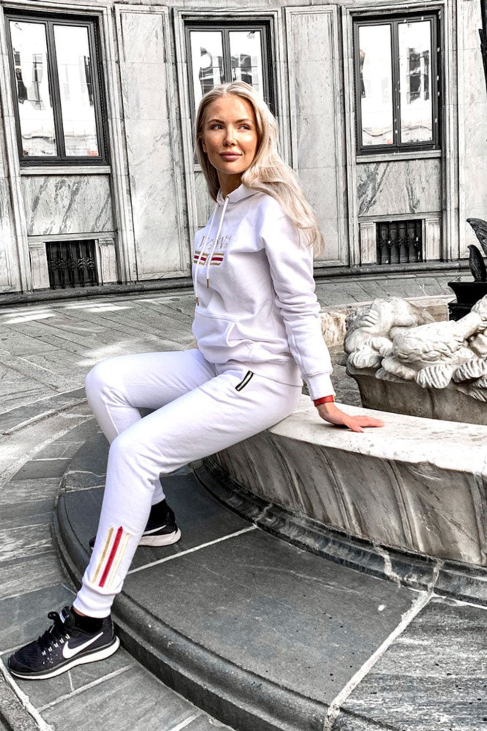 The model sits by a fountain in town in the white hoodie and white joggers with a gold stripe.