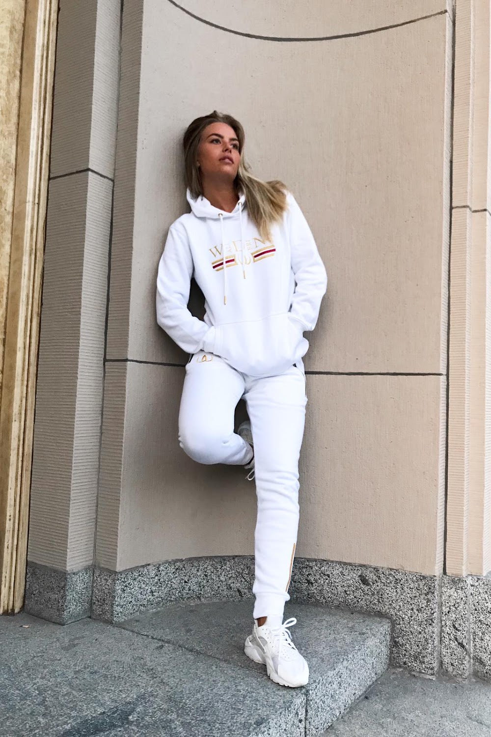 Against the house wall the model stands in a white hoodie and white joggers, worn with white sneakers.