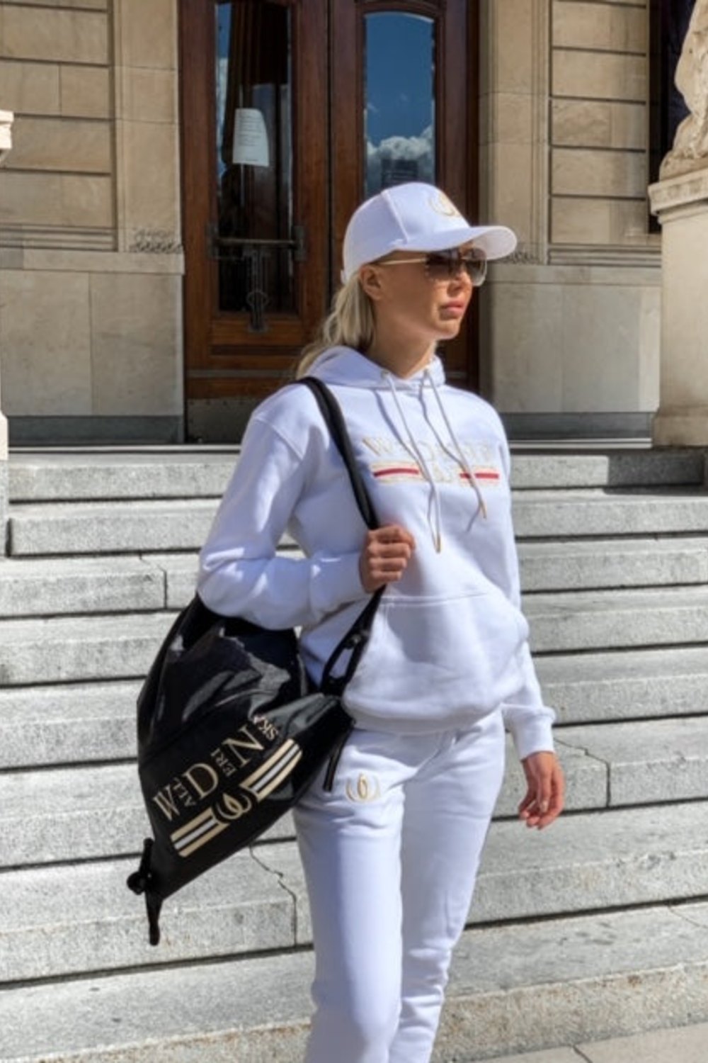 The joggers are worn with a white hoodie and cap, with a black backpack with a gold logo over the shoulder.