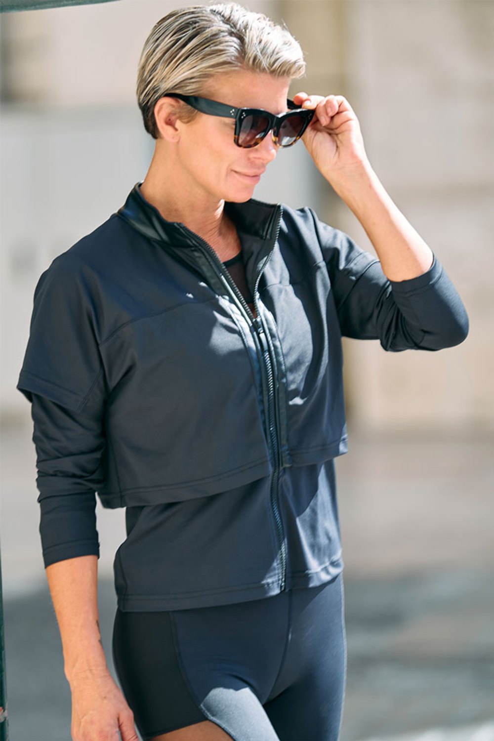 Half-open zip on the black jacket, while the model lifts her sunglasses.