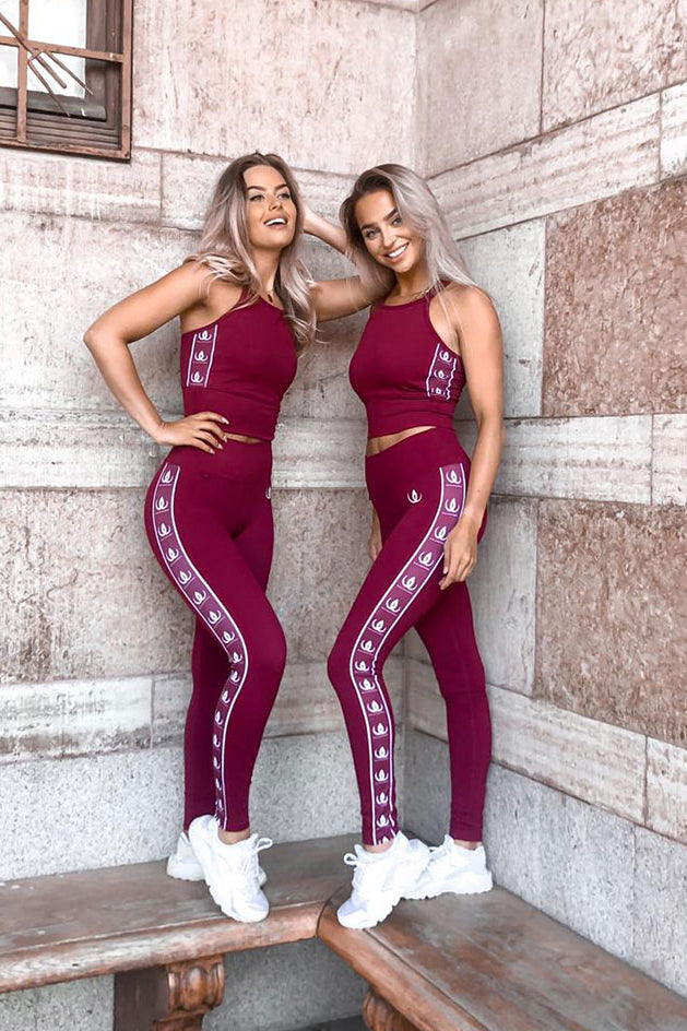 Two models leaning against a stone wall in wine red crop tops and tights with white logo bands.