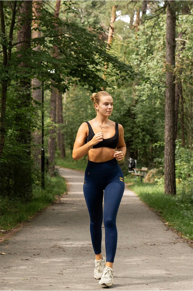 Running on a forest track in navy tights with gold stripes and a black sports bra.